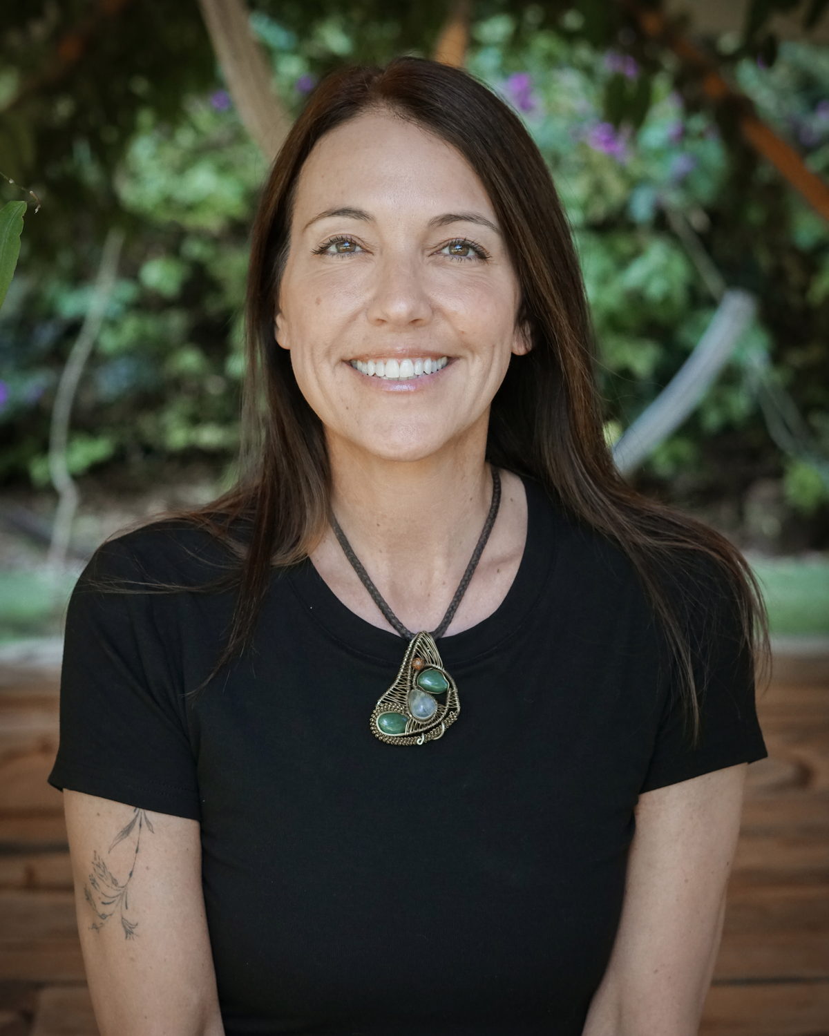 Michelle Gale, smiling, in a black shirt with a stone pendant, trees behind her